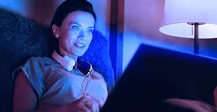Person working on laptop with blue light glow on face next to skincare products like vitamin C serum and tinted sunscreen